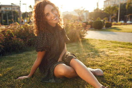 adorable curly red-haired girl with freckles in a dress enjoys good weather sitting on the lawnの写真素材