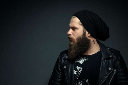 portrait of a handsome bearded man in a leather jacket and hat looking to the side.