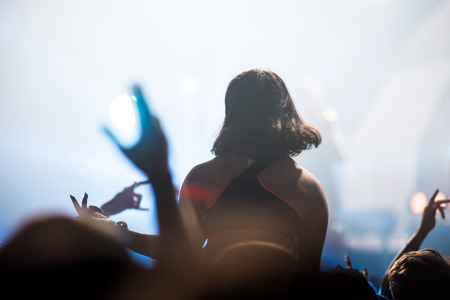 Rear view of crowd with arms outstretched at concert. Bright stage lights in the background