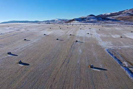 Aerial view of snow-covered field dotted with hay bales under a clear blue sky in a mountainous region during winterの写真素材