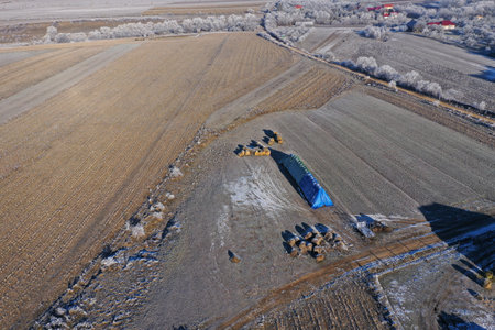 Aerial view of rural landscape showing farm fields, icy terrain, and distant village during winter morning by droneの写真素材