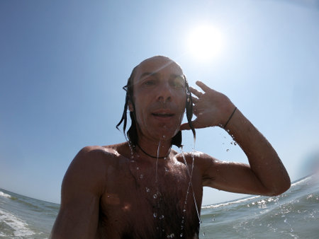 Handsome guy with long hair having fun at beach, sea waves splashing on his bodyの写真素材