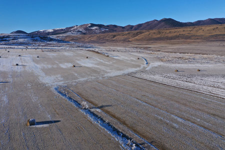 Aerial view of snow-covered field dotted with hay bales under a clear blue sky in a mountainous region during winterの写真素材