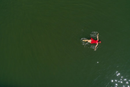 Aerial view of a woman in a red dress swimming in a calm, dark body of water. The sunlight is reflecting on the waterの写真素材