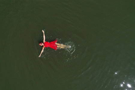 Aerial view of a woman in a red dress swimming in a calm, dark body of water. The sunlight is reflecting on the waterの写真素材