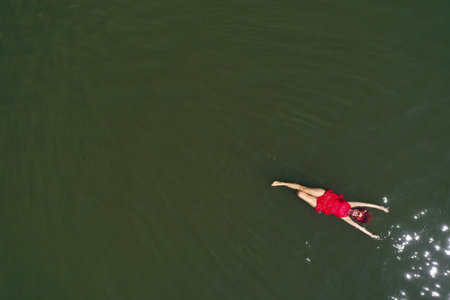 Aerial view of a woman in a red dress swimming in a calm, dark body of water. The sunlight is reflecting on the waterの写真素材