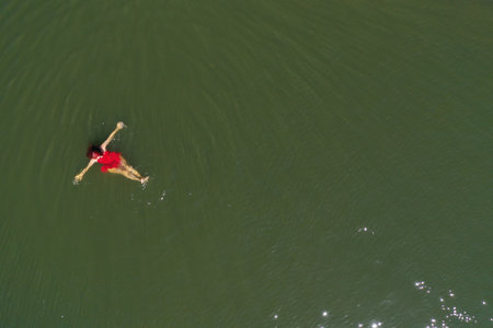 Aerial view of gorgeous woman in red dress floating on calm water by droneの写真素材