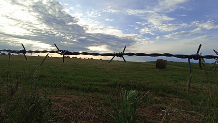 A peaceful landscape featuring a barbed wire fence in a green pasture, surrounded by hay bales and a beautiful sunset sky. Perfect for rural themes and serene outdoor settings.の写真素材