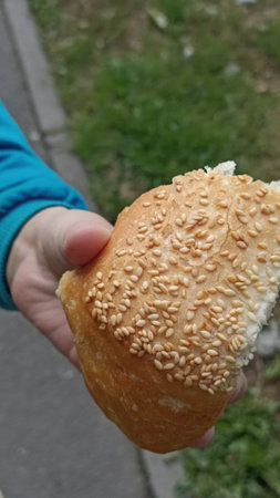 A hand holds a piece of sesame seed-covered bread outdoors, highlighting a fresh-baked snack. Ideal...