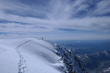 A lone hiker, warmly dressed in winter gear, stands at the summit of a snow-covered mountain,...