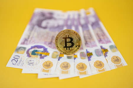 Gold Bitcoin over british pound notes on yellow background with selective focus. Cryptocurrency exchange, tax, regulation in the UK conceptの写真素材