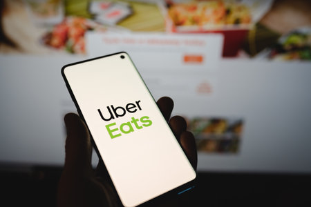Man holding a smartphone with Uber Eats logo in front of the official website. Popular food delivery service on dark blurred backgroundのeditorial素材