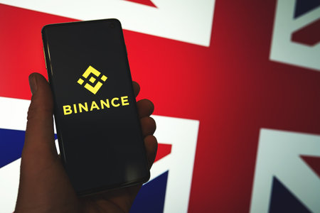 Binance app logo on smartphone in hand with the blurred British Union Jack flag background. Binance...