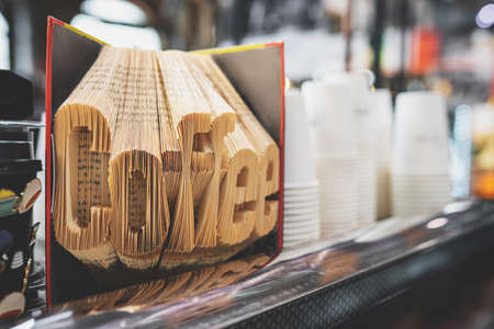 Word Coffee with folded book pages. Book sculpture decoration in a cozy coffee shop interior. White paper cups on the background. Toned image with selective focus. Coffee takeaway business conceptの写真素材