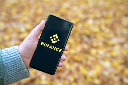 Binance app logo on smartphone in womans hand with blurred yellow Autumn foliage background. Cryptocurrency exchange, trading platform. Warsaw, Poland - October 19, 2021のeditorial素材