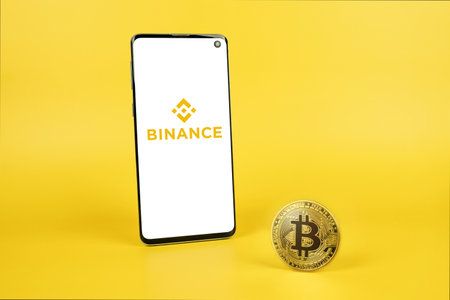 Binance logo on phone screen with Bitcoin cryptocurrency coin on yellow background. Cryptocurrency exchange, trading platform. Swansea, UK - March 31, 2021.のeditorial素材