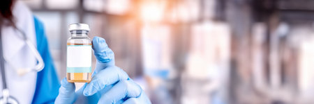 Banner with Doctor's hand holding a vaccine vial with white label mock-up for pharmaceutical ads, healthcare education, and vaccination awareness. Blurred hospital background with copy space for text.の素材