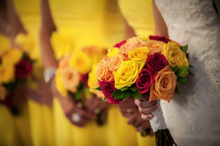 Beautiful bride holding red, yellow, orange bouquet with bridesmaids in the background.の写真素材