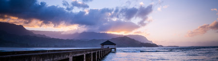 Sunset at the beach with a pier in the foreground and mountains in the backgroundの写真素材