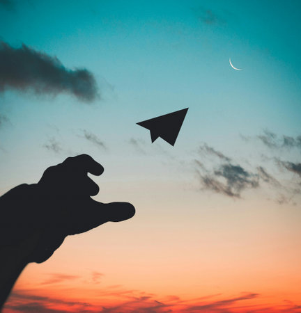 Silhouette of hand holding paper plane with sunset sky background.