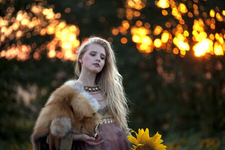 Portrait of a young beautiful girl with long blond hair in a beautiful dress in antique style on a background field of sunflowers at sunsetの写真素材