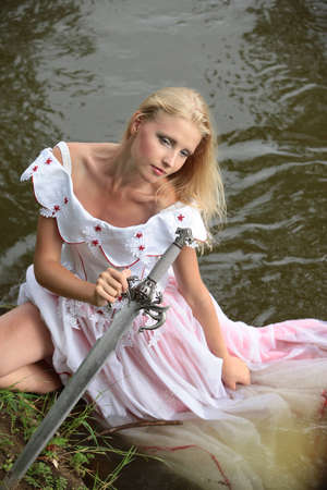 portrait of a beautiful young blonde in a pink dress with a sword in hand, standing in the river in summer day on background green overgrown bushes coastの写真素材