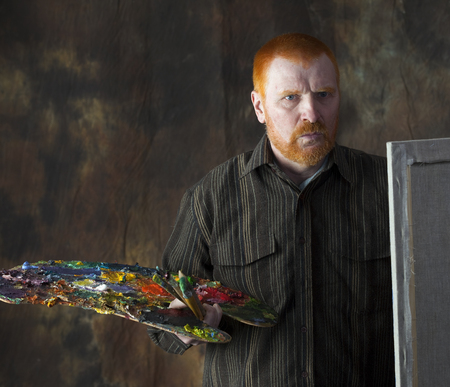 close-up portrait of an adult male artist with red hair and a beard at work studio on dark backgroundの写真素材