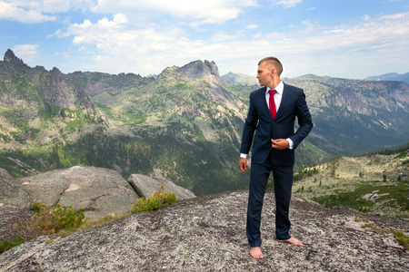 portrait of a young man in a business suit at the top of the mountain on the background of mountains and blue sky on a sunny summer dayの写真素材