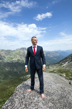 portrait of a young man in a business suit at the top of the mountain on the background of mountains and blue sky on a sunny summer dayの写真素材