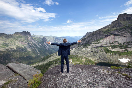 portrait of a young man in a business suit at the top of the mountain on the background of mountains and blue sky on a sunny summer dayの写真素材