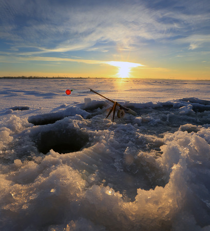 close-up drill, fishing rod near the hole on the ice in winter river at sunsetの写真素材