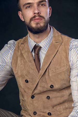 closeup portrait of the beautiful charismatic young man with a beard wearing pants, shirt, vest and tie on a dark background studioの写真素材