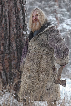 Portrait of adult man with long hair and a beard in a sheepskin coat, with an ax in the winter...