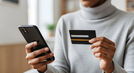 A person is holding a smartphone and a credit card, preparing to make an online payment or transaction. The focus is on the hands and the devices, emphasizing the convenience and pの素材