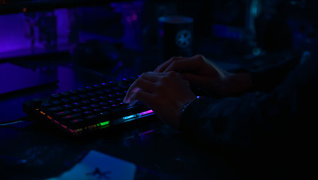 Close-up of hands typing on a backlit mechanical keyboard bathed in blue and purple neon light. The moody, high-tech setup conveys focus and gaming culture, suitable for tech gear, esports, and digital workstation advertising.の素材