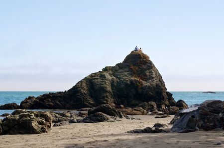 Two people sitting atop a very large rock on a California beach.の写真素材