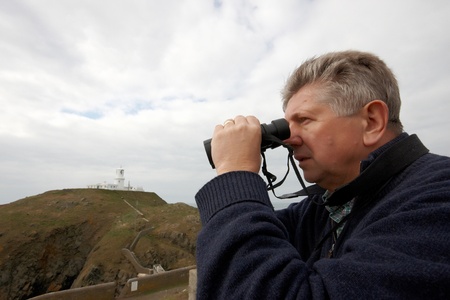 Mature man looking through binoculars with light house in the distance