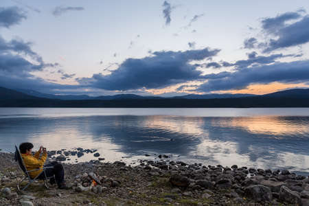 Lofoten, Norway- September 12 2020: Tourist sitting near fire on autumn evening and enjoying the view on a fjord in Norway. Taking pictures of a passing duck on a mobile phoneのeditorial素材