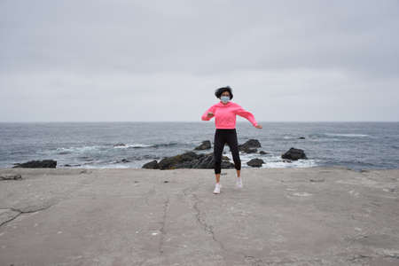 Woman wearing a mask exercises in front of the sea. New normality. Sports session
