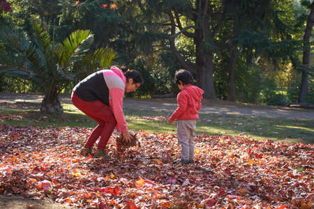 Mother picks up dry leaves from the ground in front of her child. Woman with child in the park....