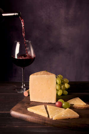 A piece of Manchego Cheese from Spain with some green and red grapes and a glass and bottle of Spanish red wine at the background in a dark food photography styleの写真素材