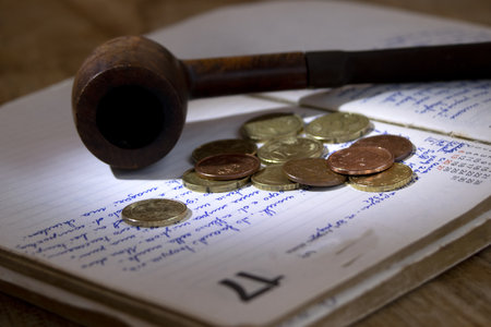 An old wooden pipe and scattered coins on a vintage diary, evoking notions of thrift, personal history, and simplicityの写真素材