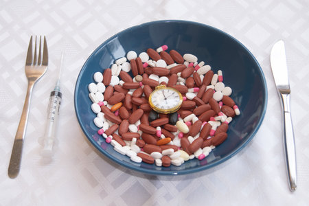 a blue bowl filled with various pills and a golden pocket watch, flanked by a fork, knife, and syringe. time for medicationの写真素材