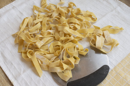 a pile of golden fresh homemade fettuccine or tagliatelle pasta, lightly dusted with flour, resting on a rustic white kitchen towel with a pasta cutter underneath. raw italian food preparation.の写真素材