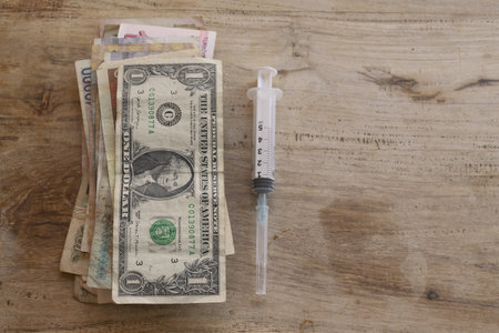concept of high healthcare expenses and drug costs with a stack of dollar bills and other currency notes next to a medical syringe on a rustic wooden backgroundの写真素材