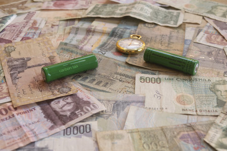 green batteries labeled carbon tax placed on collection of old international banknotes with pocket watch symbolizing environmental taxation and energy transition costsの写真素材