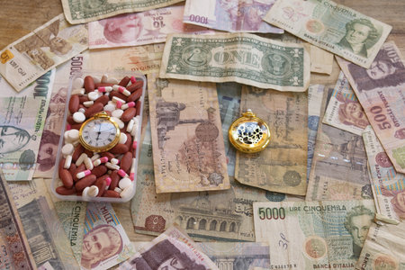 assorted pills and gold pocket watch on various international banknotes symbolizing time, health and money in global healthcare economyの写真素材