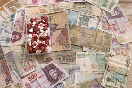 colorful pills, tablets and capsules in transparent plastic container placed on scattered international money banknotes, expensive healthcare and medication cost conceptの写真素材