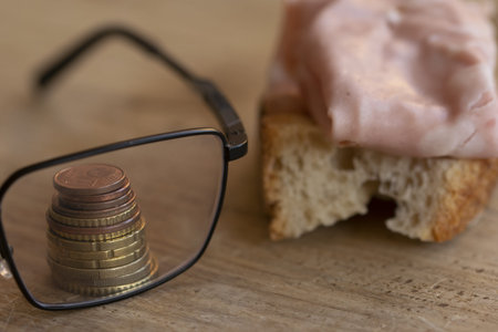 A stack of euro coins seen through eyeglasses next to a mortadella bread slice on a wooden tableの写真素材