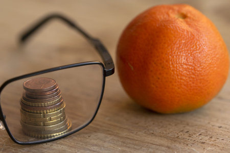 financial planning concept showing stacked euro coins viewed through reading glasses beside fresh orange on wooden tableの写真素材